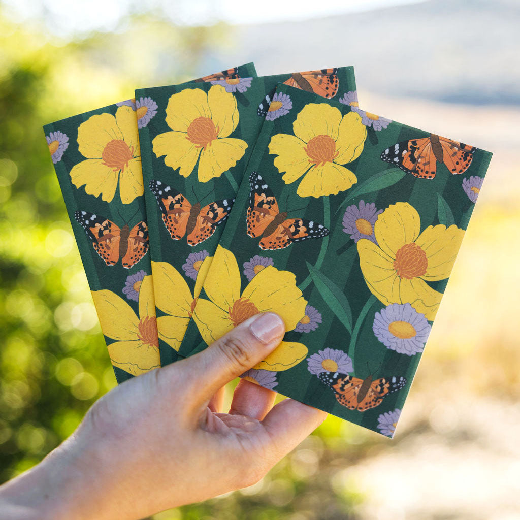Three green cards with yellow flowers and butterflies held by a hand against a blurred natural background.