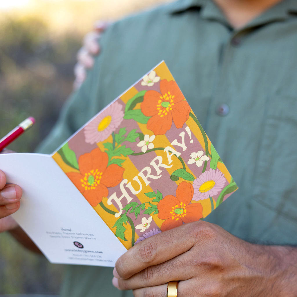 Person holding a card with floral design and 'Hurray!' text, outdoors golden hour