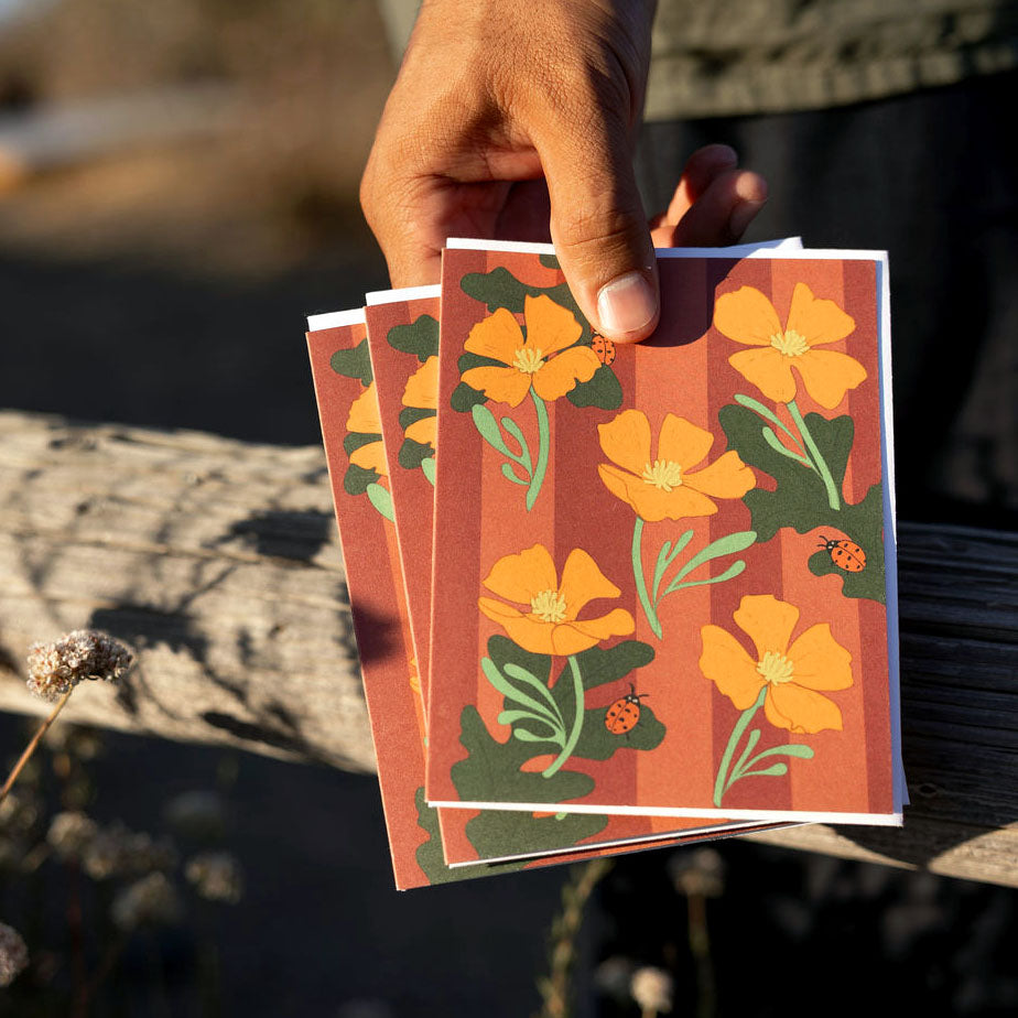 Hand holding a stack of floral greeting cards with orange poppies and burgundy stripes on a natural background.